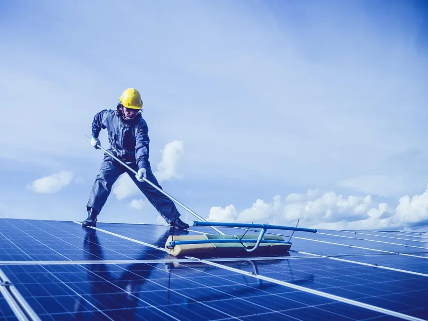 Technician in a hard hat cleaning a large array of solar panels under a bright sky