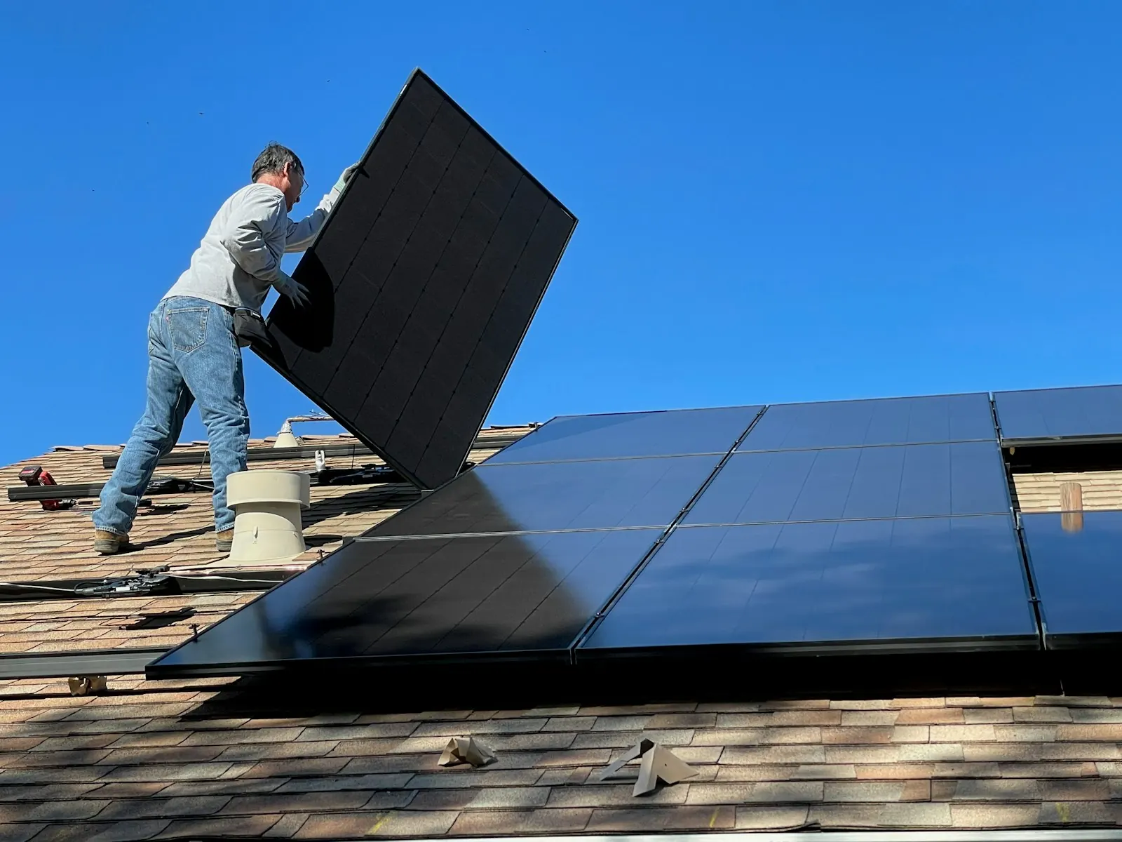 Installer carrying a large black solar panel across a shingle roof under a blue sky