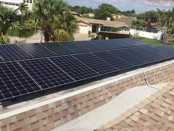 Rows of black solar panels on a light shingle roof surrounded by palm trees