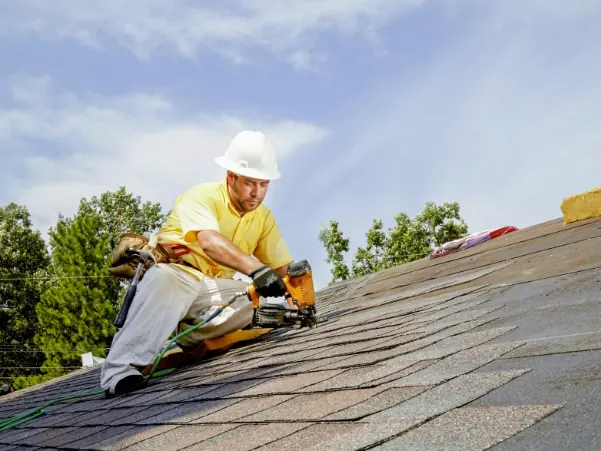 Roofer in a white hard hat using a nail gun on a sloped shingle roof